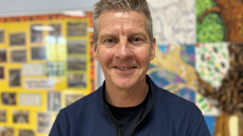 Steve Cram pictured smiling against a colourful paper display. He has grey hair and is wearing a dark blue round-neck shirt underneath a dark blue jacket.