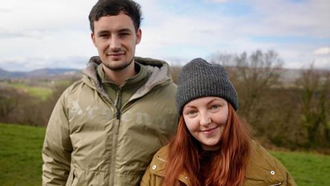 Jade and Jamie are standing in a meadow, side by side, Jade is wearing a beanie hat and Jamie a green coat.