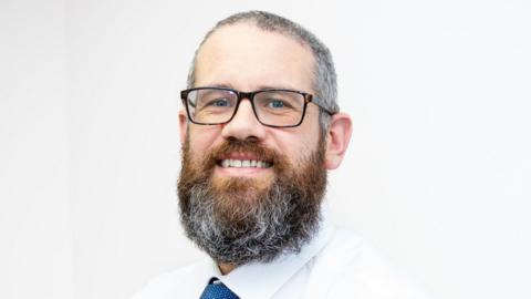 An image showing the head and shoulders of a man with a grey beard who is wearing glasses and smiling for the photograph. He is also wearing a white shirt and a blue tie and is standing in front of a white background.