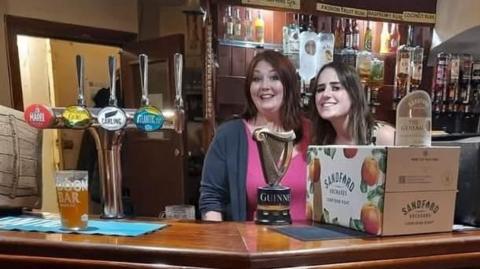 Two women behind a bar smiling at the camera with a pint on the bar