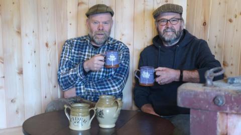 Tim and Steve sit at a wooden table holding mugs. Both are dressed in flat caps, Tim on the left is wearing a chequered shirt and Steve is wearing a hoodie. Steve on the right is also wearing glasses. Two jugs that say the word cider sit in the middle of the table.