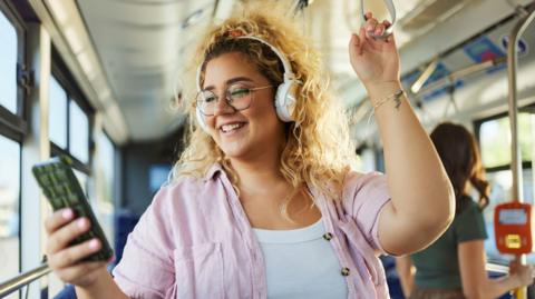 A woman with curly, blonde hair is listening to music and smiling while on a bus. She's wearing a pink shirt.