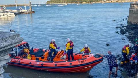 An orange RNLI rib sits close to rocks at the bottom of a sea wall where the tide is starting to come in. A boy is laying on a stretcher on the rocks in shallow water while three rescuers lean over him. On the rib are three other RNLI crew members in yellow and red jackets and white helmets. At the top of the wall is a stone balustrade. It's a sunny day, the water is calm and there are some small boats moored in the distance.