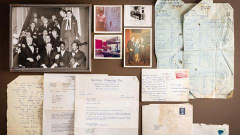 A picture of a number of letters on a table. They are pictured to the bottom of the image and spread out to the right side. In the top left corner, there are five photographs.