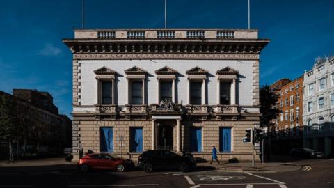 A white and brown building with columns at the sides of the windows. Cars are parked outside. A person wearing a blue coat is walking by. The sky is blue with minimal clouds. 