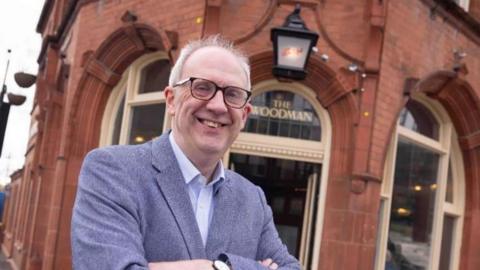 A photograph of a man wearing glasses stood outside a traditional pub with "The Woodman" written on glass above the door behind him. He has his arms crossed and is wearing a suit jacket and shirt. 