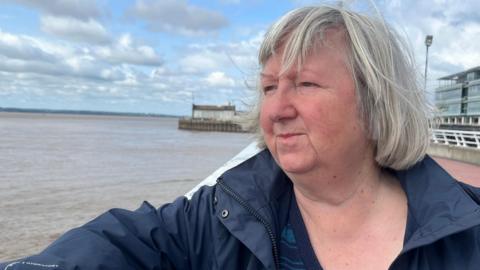 A woman with light-grey hair in a bob wears a blue jacket and stands on a promenade staring across the grey-brown waters of a wide river estuary. White railings and a large glass building can be seen to her right. The sky is blue and dotted with white clouds.