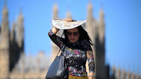 A woman in bright sunlight shades her face with a patterned paper fan