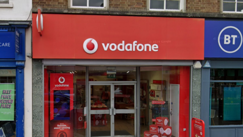 The outside of the Vodafone shop on the High Street in Newmarket. It has a red sign saying "vodafone" over the door and glass windows and doors. 