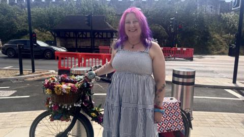 A woman with pink and purple ombre hair and pale skin stands in front of a cordoned off cycle lane with her bicycle. She is wearing a navy and white stripe sundress, and blue and red bangles on her wrist. Her bicycle is black with a basket on the front and fake flowers and fairy lights twined around it.