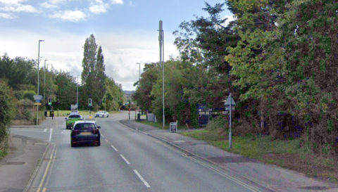 A road through a residential area leads down to some traffic lights, which are on green as a white car cuts across the junction coming from the opposite direction. There is a side road to the left shortly before the junction, and a right turning to a sports club. There are a lot of trees surrounding the roads.