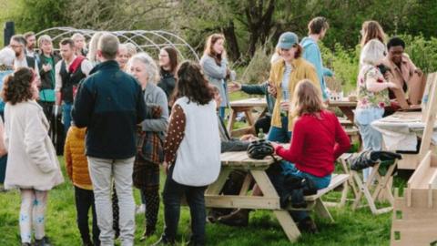 Knowle West Spring Celebration - shows groups of people standing and sitting on benches, eating and drinking together in a field