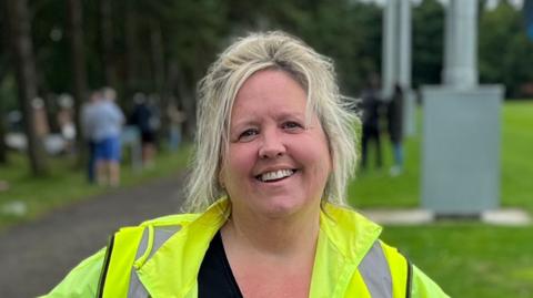 A woman with light blonde hair smiles into the camera. She is wearing a yellow high visibility jacket and a black top. The background is blurred, but you can make out people standing beside trees and grass. 