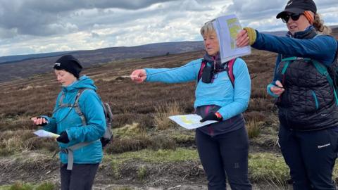 Three women in the holding maps and compasses pointing ahead on the fells.