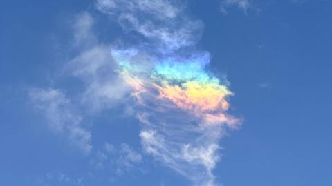 A blue sky has an unusual cloud formation in the middle with white, blue, yellow, red and green tones. It looks like a partial rainbow has formed in the clouds.