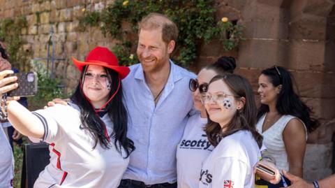 Prince Harry, wearing a light blue shirt, smiles for a photo with three young women. The woman taking the selfie is wearing an England football shirt and a red cowboy hat.