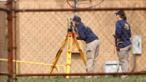 Two FBI agents gather evidence at the scene of the attempted assassination on Trump. One woman looks into a surveying device as another looks on.