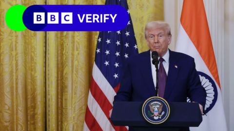 President Trump standing in front of a gold curtain and the flags of India and the USA. He is behind a blue lectern featuring the presidential seal. The BBC Verify logo is in the top left of the image.