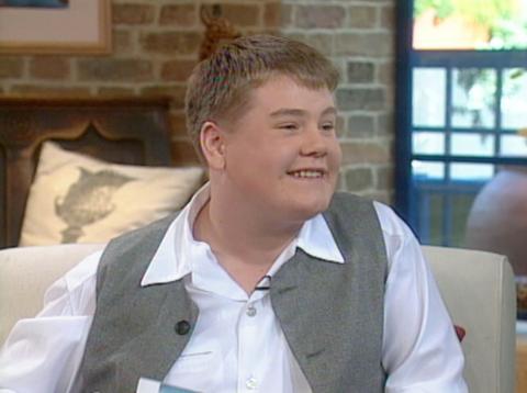 A young James Corden in a waistcoat, on a studio couch, smiling.