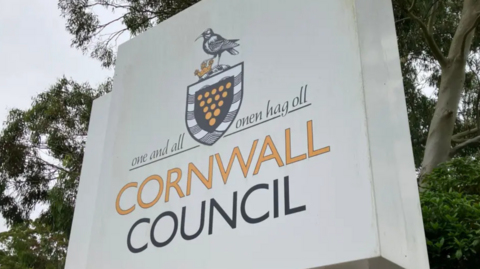 Cornwall Council sign on a white background. There are trees in the background. The words are written in black and orange colours. 