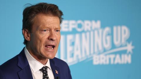 Richard Tice in a dark suit, wearing a union flag button on his lapel. He is standing behind a large turquoise blue sign with white writing which reads: Reform cleaning up Britain 