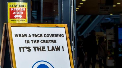 A business in a shopping centre displays a sign asking people to wear a face covering during the Covid pandemic. A further sign reminds people to "stay safe" and follow social distancing rules and keep two metres apart.