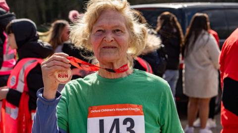 Eileen Williams holding a medal after one of the OX5 Runs. She is smiling. She wears a top with a sticker with the number 143 and Oxford Children's Hospital Charity. There are other people behind her, some of whom are event marshals in pink high vis jackets.