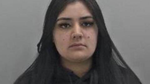 Sanaa Shahzad is a young woman who has long straight, dark hair and is seen wearing a black jumper. She has brown eyes and a mole on each cheek. 