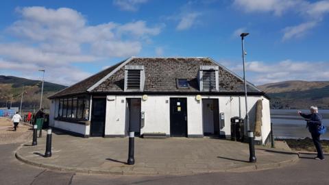 A large white and black building that is a public toilet. It is pictured on a sunny day, with hills and a loch visible behind it
