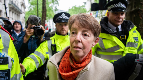 Former Post Office chief executive Paula Vennells, wearing an orange scarf and tan jacket, flanked by police in hi-vis jackets.