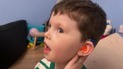 A young boy with short brown hair turns his head to show his orange and blue hearing aid in his ear. He is wearing a green and white t-shirt and is standing in a blue painted room with toys on the floor.