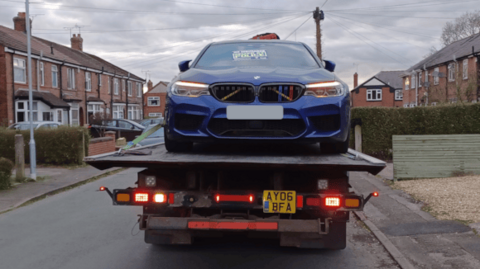 A blue sports car is hauled away on a truck through a residential area. 