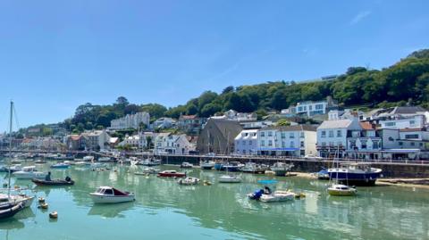 A picture of St Aubin. There is a number of houses and shops in the distance. There is boats and a large body of water in the image.