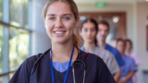 Health professionals standing in a line in a hospital corridor, with a woman at the front smiling at the camera.
