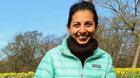 Dr Sanjana Kochcar – known as Sanj - smiles at the camera sitting in a field filled with daffodils, wearing a mint green bubble coat, with brown turtleneck jumper.