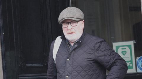 A 62-year-old man with a black padded jacket, a green tweed cap and glasses outside a court building