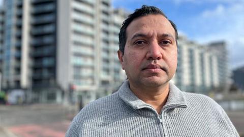 A man with short black hair in a close-up shot in front of a high-rise block of flats. 