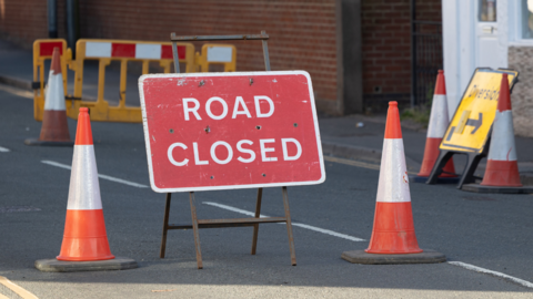 A red sign with "ROAD CLOSED" written in capital letters in white in the middle of a road. Two orange and white traffic cones sit to either side of the sign. There are barriers and traffic cones in the background, as well as a yellow sign labelled "diversion". 