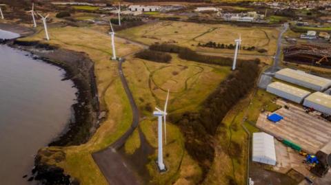 The Oldside site in Workington. The fields have several white wind turbines and it is next to the coast. Warehouses can be seen to the right.