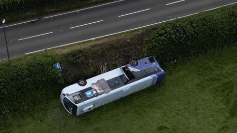 A bus overturned on grass near a main road 