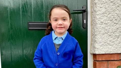 Izzy in a blue school uniform standing in front of a green door smiling at the camera. She also has a grey dress with a butterfly-shaped zip.