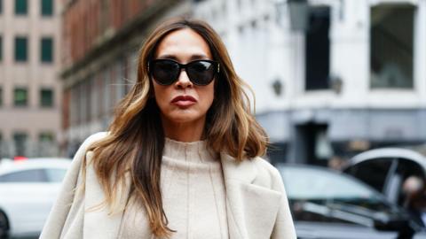 A woman with long brown hair is pictured wearing large black sunglasses and a white jumper and white coat. 