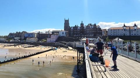 A view of Cromer from Cromer Pier. In the foreground on the right people are standing on the pier. To the left people are paddling or on the beach. In the background is the town's skyline including the Victorian Hotel De Paris.
