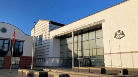 A white court building this a glass front. It has a crest in black on the right hand side and the words Isle of Man Courts of Justice above the glass. The front has a white pillar down the centre and there are two flag poles in front of the building the red Manx flag, which features a white three-legs-of-Mann.