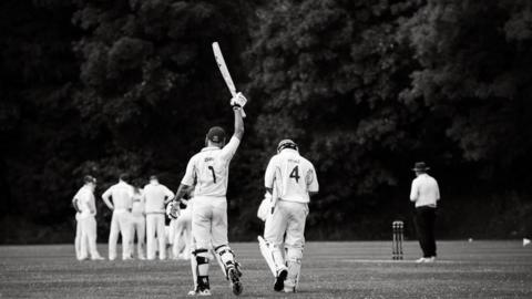 A black and white photo of players Zayn and Ayyaz going out to bat for Oxhenford's 1st XI. Zain is holding the bat above his head. Other players could be seen in the distance.