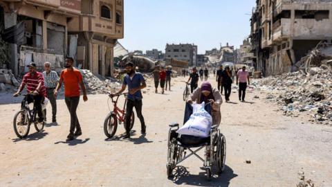 A woman pushes a wheelchair loaded with a sack of flour as she walks past rubble and damaged buildings along a street in the Tuffah district of Gaza City, in northern Gaza (8 July 2024)