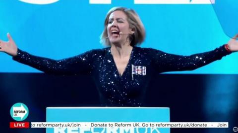 A woman with a blonde bob has both her arms out in the air and her mouth open as she is mid-singing. She is wearing a sparkly blue top and a union jack flag badge on her. She is standing on stage with a sign saying Reform UK in front of her.