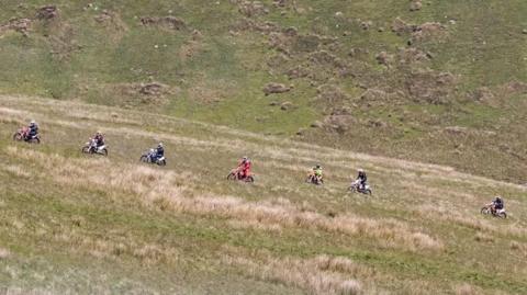 Seven people on multicoloured off-road motorcycles riding across an open stretch of green fields. 