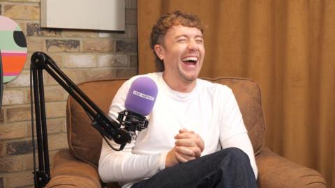 Roman Kemp sat on chair in front of a purple microphone and laughing