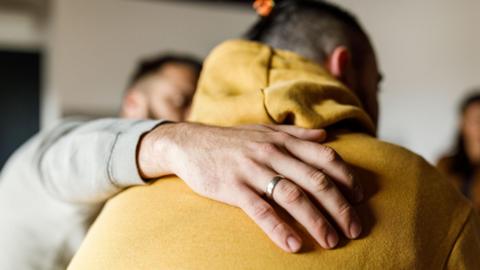 A man putting his arm around another man to comfort him. They are both in sweatshirts or hoodies. The man being comforted has his back to the camera and the other man's face is blurred.